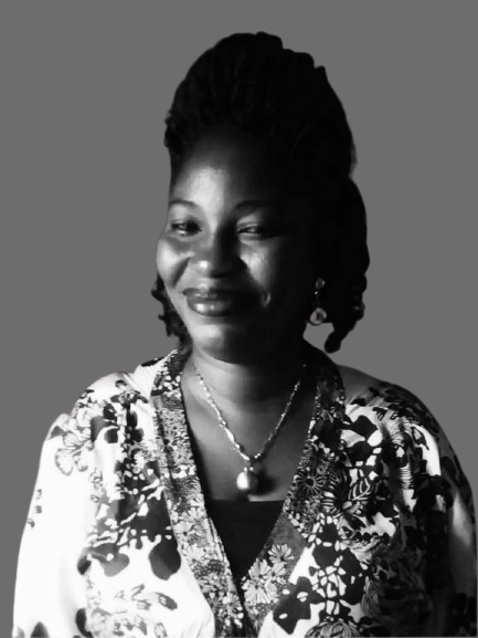 A woman with braided hair stands calmly against a plain gray background, wearing a floral-patterned black and white outfit and a necklace with a large pendant. The setting is simple and respectful, conveying a serene and dignified mood. The lower left corner contains the watermark WPS Photos.
